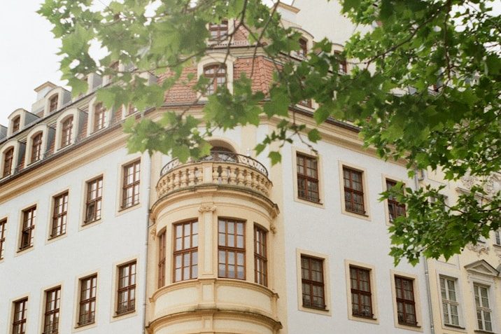 Personalvermittlung jorbis Weiße Gebäudefassade mit einer Rundbalkon und umgebenden grünen Blättern.