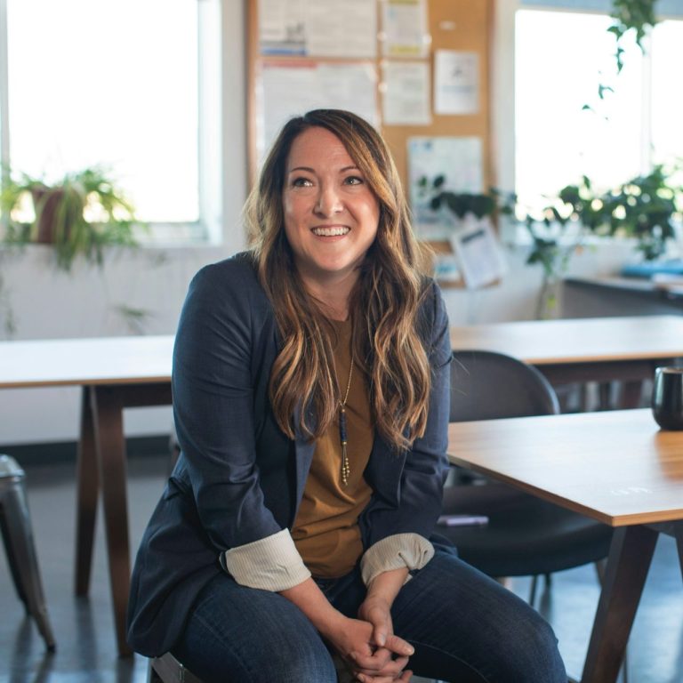 Personalberater jorbis Junge Frau im jorbis Team mit langen Haaren sitzt lächelnd an einem Tisch in einem hellen Raum.